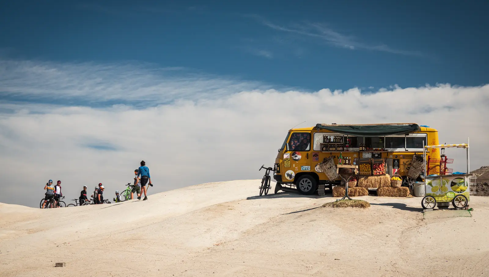 A yellow coffee van parked on a white ridge above the clouds with a group of cyclists arriving